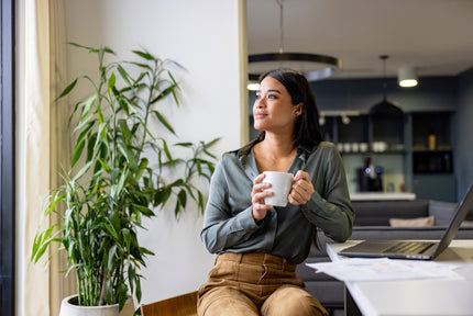 Frau sitzt mit Kaffee vor Laptop und schaut aus dem Fenster