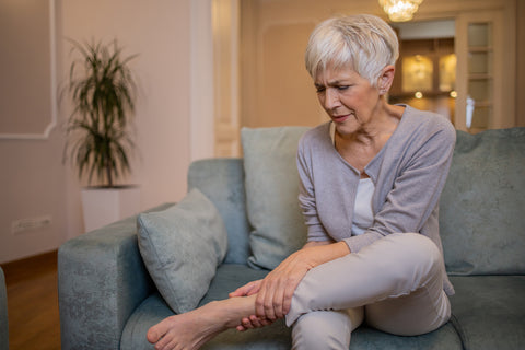 Erfahrung Meno-Balance-Kur: Frau sitzt auf dem Sofa und befühlt ihren schmerzhaften Knöchel