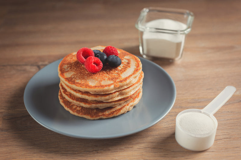 Protein Pancake Rezepte: Ein Teller mit Stapel Pancakes und Obst als Topping, daneben Messlöffel mit Proteinpulver