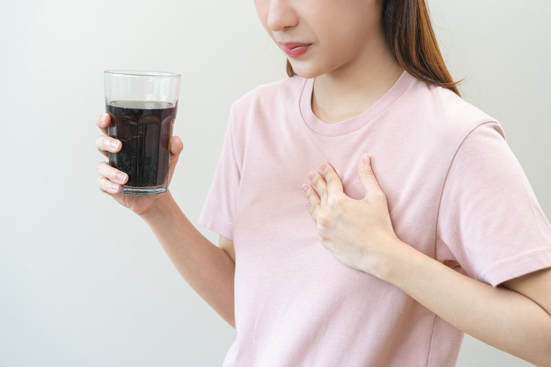 Mittel gegen Sodbrennen: Frau hält ein Glas Cola in der einen Hand, mit der anderen fasst sie sich an die Brust