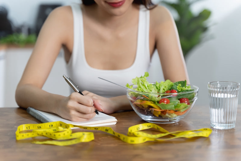 Frau schreibt ein Ernährungstagebuch, daneben ein Salat und ein Maßband