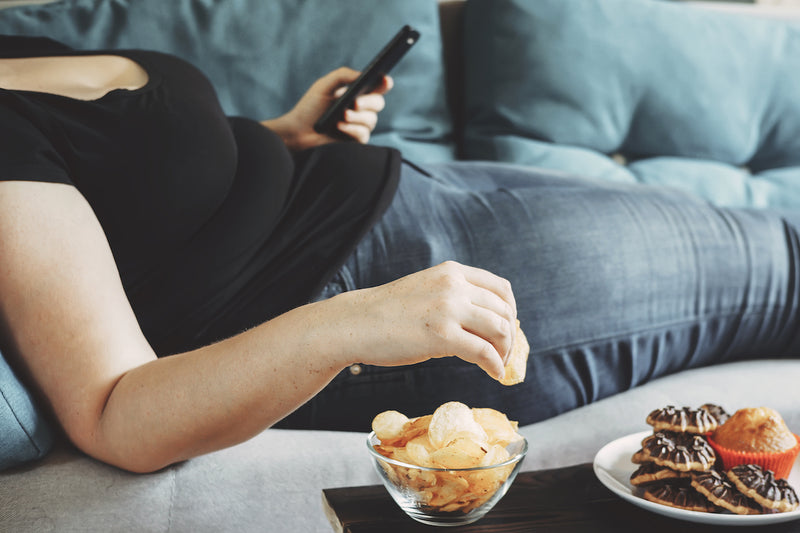 Person liegt mit Fernbedienung auf der Couch, neben ihr stehen Chips und Süßigkeiten