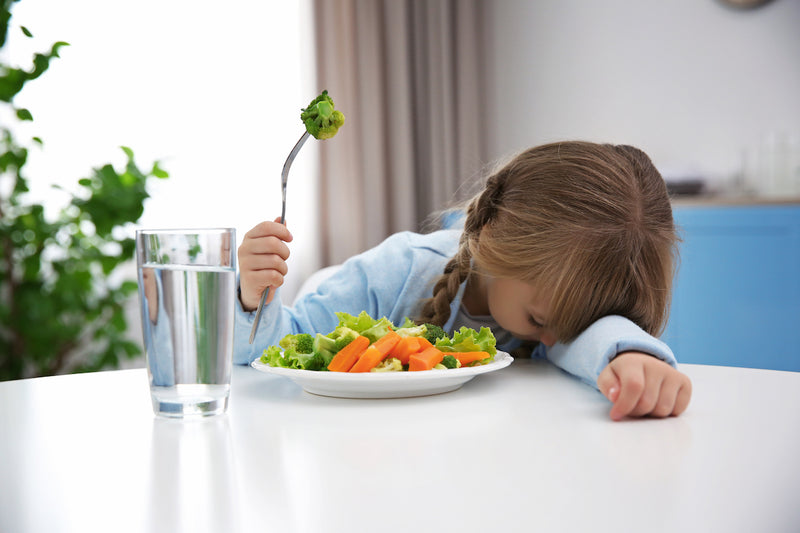 Kind mit Gemüse auf Gabel legt den Kopf auf dem Tisch ab