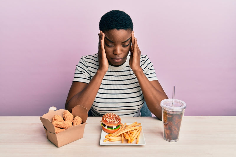 Frau sitzt vor Fast Food und hält sich den Kopf vor Schmerzen
