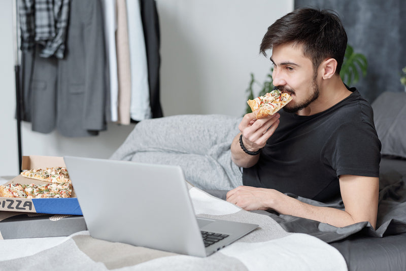 Mann isst Pizza im Bett vor dem Laptop