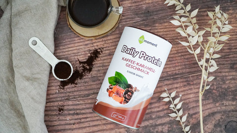 Das VitaMoment Daily Protein Kaffee-Karamell auf einem Tisch mit einem Löffel Pulver und einer Kaffeetasse