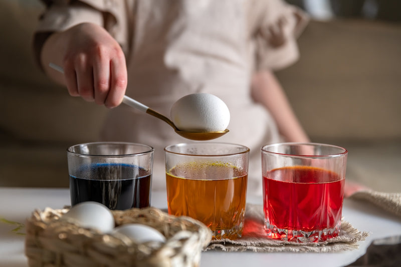 Ostereier natürlich färben: Kind gibt Ei auf einem Löffel in farbiges Wasser in Gläsern