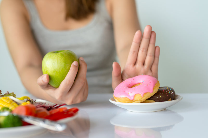 Frau hält Apfel in der Hand und schiebt Teller mit Donuts weg
