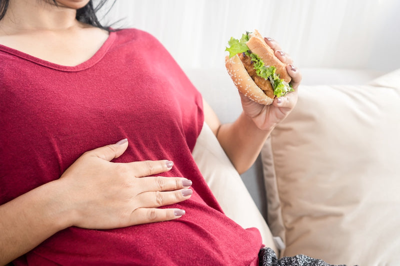 Mittel gegen Sodbrennen: Frau hält einen Burger in der Hand und fasst sich mit der anderen Hand an den Bauch