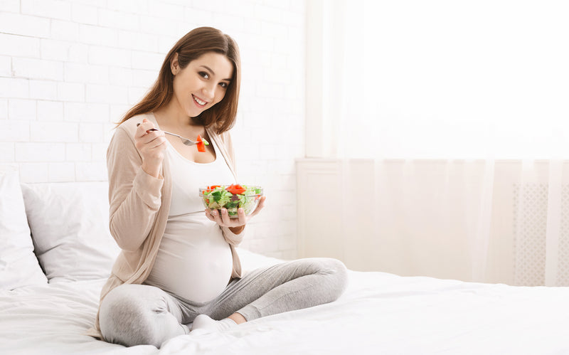 Nährstoffe in der Schwangerschaft: Schwangere Frau sitzt auf dem Bett und isst einen Salat
